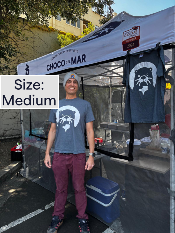 A person standing with a shirt that has Cleo Otter the mascot of Choco Del Mar on it. Size: Medium